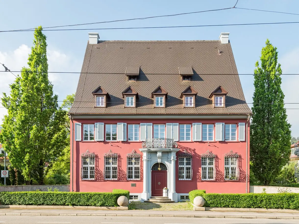 Barockes Baudenkmal mit rot verputzter Fassade, Fensterläden, verzierten Fenstergittern, Säulenportal und Walmdach mit Gauben