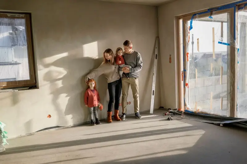 Junge Familie mit zwei kleinen Kindern steht in einem Raum ihres Hauses während der Sanierung: verputzte Wände, Estrich, abgeklebte neue Fenster