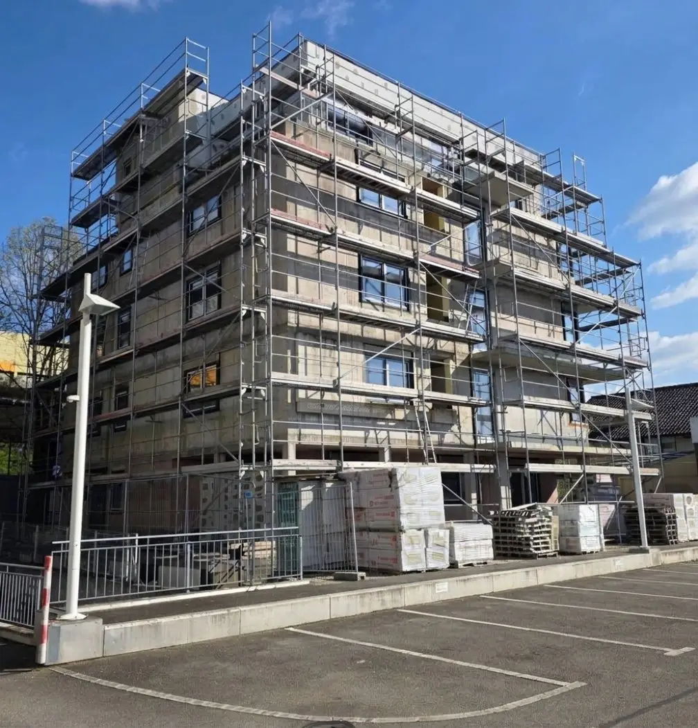Mehrfamilienhaus im Rohbau mit Gerüst, die Fassade wird gedämmt; davor Paletten mit Dämmplatten und ein Parkplatz